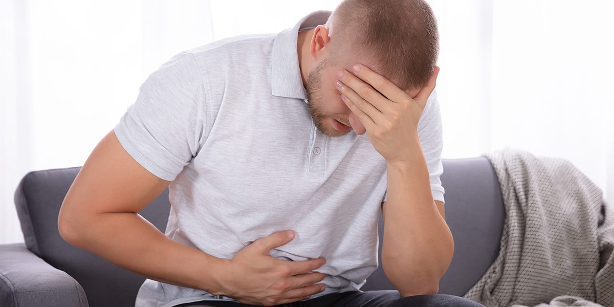 Sad Young Man Sitting On Sofa Suffering From Stomach Pain